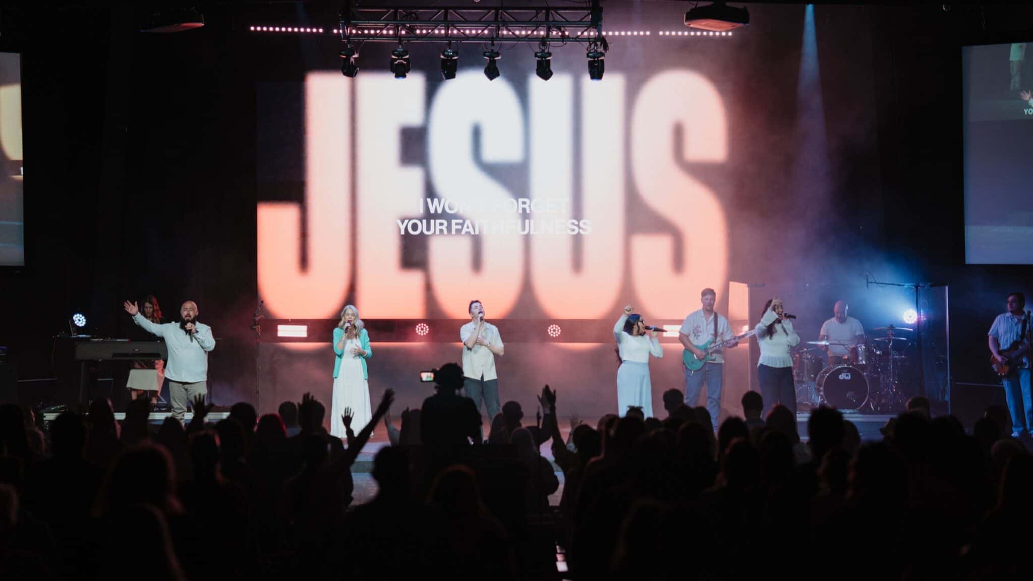 Worship leaders on stage before a large JESUS screen and crowd